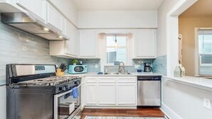 Una cocina con gabinetes blancos, estufa, microondas, fregadero y ventana.