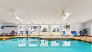 An indoor swimming pool with clear glass walls, blue lounge chairs, and ceiling fans.