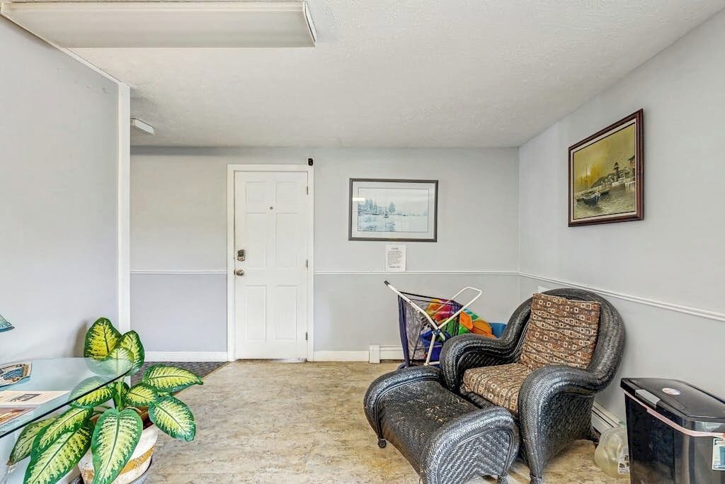 A room with a door, a framed picture, a wicker chair, a trash bin, and a plant.