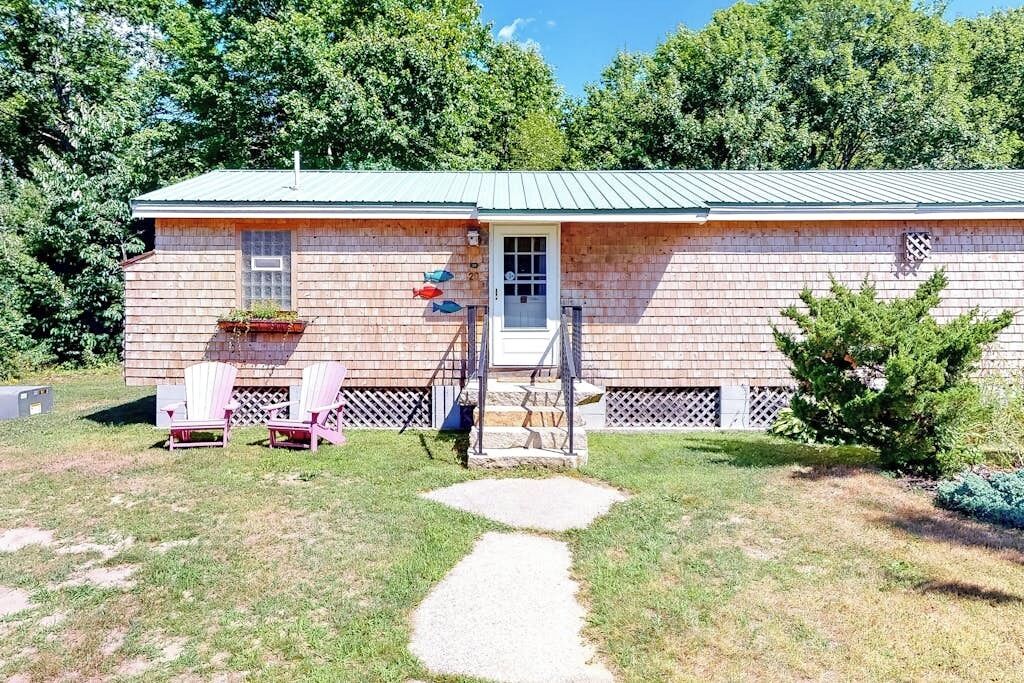 A single-story house with a metal roof, a front door, and a small porch area with two Adirondack chairs.
