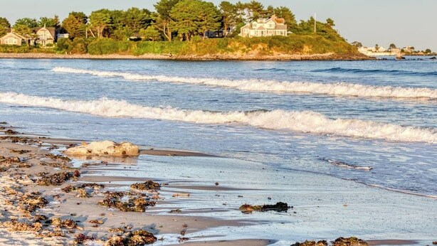 A beach with waves, a sandy shore, and a house in the background.