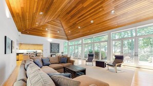 Un amplio espacio habitable con techo de madera, ventanas grandes, un sofá modular y una cocina moderna al fondo.
