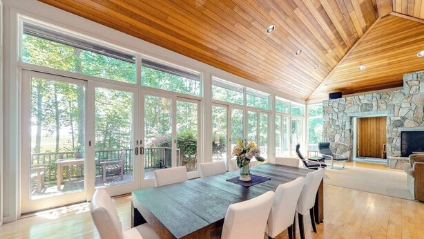Un comedor espacioso con una gran mesa de madera y sillas blancas, una chimenea de piedra y ventanas amplias que ofrecen vistas al exterior.