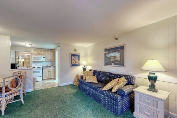 A room with a blue patterned sofa, a small white nightstand with a lamp, a dining area with a chair and table, and a kitchen in the background.