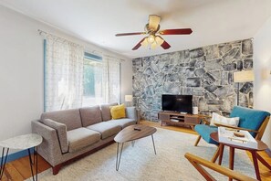 A living room with a sofa, coffee table, and armchair, a stone wall, a television, and a ceiling fan.