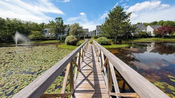 Een houten brug over een vijver met waterlelies, omringd door huizen en bomen.