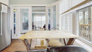 Un comedor con una mesa de madera y sillas metálicas, una cocina con armarios blancos y vistas al exterior a través de amplios ventanales.