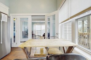 A dining area with a wooden table and metal chairs, a kitchen with white cabinets, and a view of the outdoors through large windows. - Charming condo with outdoor pool, private washer/dryer, partial estuary view (Wells)