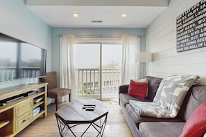 A living room with a flat-screen TV, a wooden coffee table, a sofa with cushions, and a balcony with a view. - Charming condo with outdoor pool, private washer/dryer, partial estuary view (Wells)
