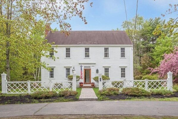 A two-story white house with a front porch, a flag, and a white picket fence. - Idyllic Retreat near the Beach with Fast WiFi, Two Living Areas & Full Kitchen (Kennebunk)