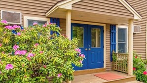 A house with a blue front door, a porch with a wooden railing, and a large flowering bush in front. - Lovely townhome near golf with gas grill, patio, high-speed WiFi, & full kitchen (Ogunquit)