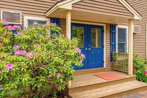 Ein Haus mit einer blauen Haustür, einer Veranda mit Holzgeländer und einem üppigen, blühenden Busch davor.