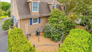 A two-story residential house with a sloped roof, a visible attic, and a small garden area with a trellis structure. - Lovely townhome near golf with gas grill, patio, high-speed WiFi, & full kitchen (Ogunquit)