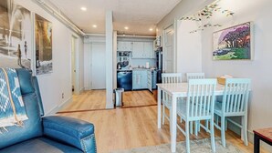 A modern living room with a sofa, dining table, and chairs, a kitchen in the background, and artwork on the walls. - Ocean-view condo overlooking Ogunquit Beach with balcony, pool & tennis (Ogunquit)