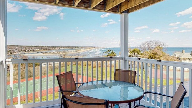 A covered outdoor area with a table and chairs, overlooking a sports court and a coastal view. - Ocean-view condo overlooking Ogunquit Beach with balcony, pool & tennis (Ogunquit)