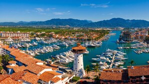 Una marina con numerose imbarcazioni, un faro ben visibile e una cittadina costiera sullo sfondo con edifici e montagne.