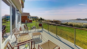 Eine Terrasse mit Sitzgelegenheiten im Freien, Blick auf das Meer und ein Gebäude im Hintergrund.