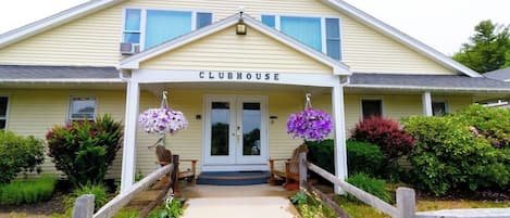 A two-story building with a porch, labeled 'CLUBHOUSE,' featuring a front entrance, a small porch seating area, and a pathway leading to the entrance.