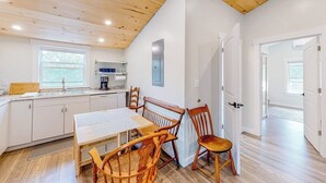 A modern kitchen with white cabinets, a wooden dining table, and chairs, a window with a view, and a door leading to another room. - Shorelands, Unit 44-3  | Condo in Kennebunk with pool and playground access (Kennebunk)