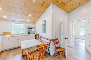 A modern kitchen with white cabinets, a wooden dining table, and chairs, a window with a view, and a door leading to another room. - Shorelands, Unit 44-3  | Condo in Kennebunk with pool and playground access (Kennebunk)
