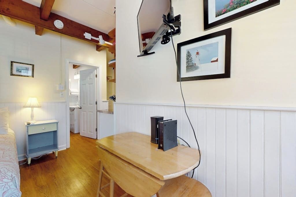 Une pièce avec un bureau en bois, un ordinateur, une lampe, un tableau encadré et une porte qui donne sur une autre pièce.