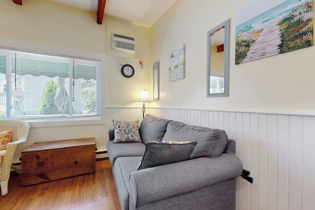A living room with a grey sofa, a wooden chest, a lamp, and a window with blinds.
