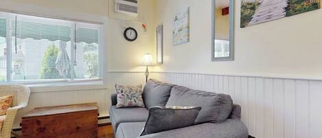 A living room with a grey sofa, a wooden chest, a lamp, and a window with blinds.