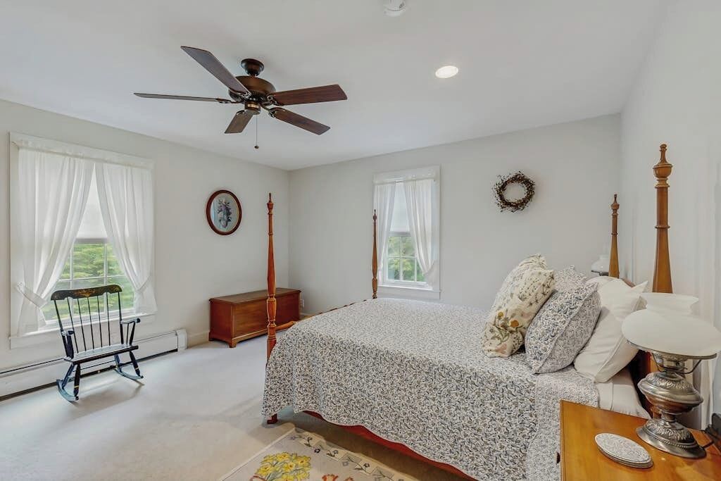 A bedroom with a four-poster bed, a ceiling fan, a wooden nightstand, and a view of the outdoors.