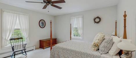 A bedroom with a four-poster bed, a ceiling fan, a wooden nightstand, and a view of the outdoors.