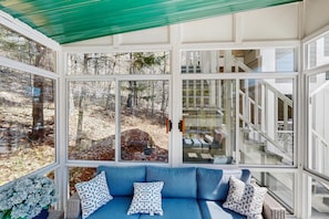 Ein Wintergarten mit blauer Couch, gemusterten Kissen und Blick auf ein bewaldetes Gebiet.