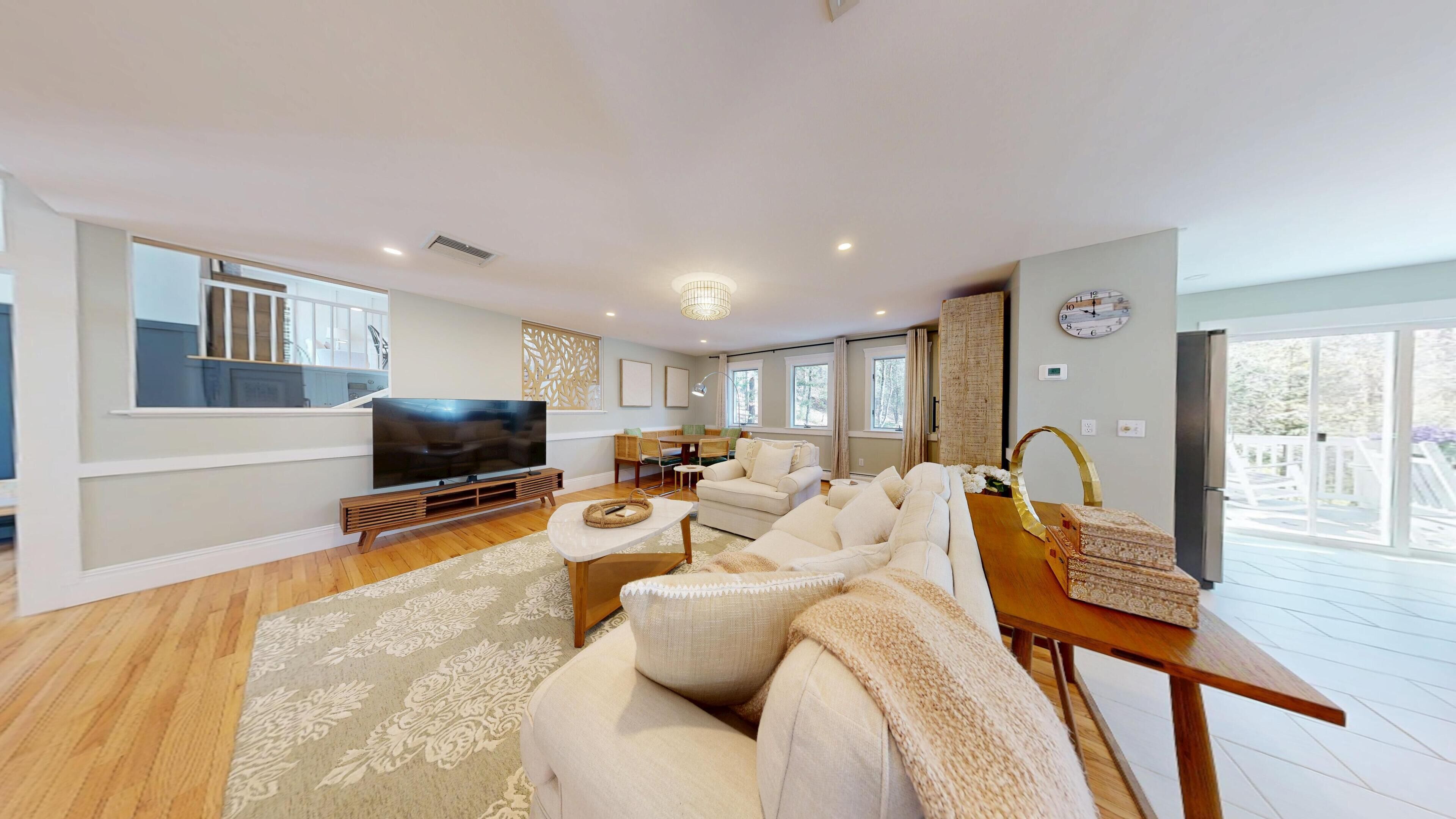 A spacious living room with a large flat-screen TV, a sofa set, a dining area, and a view of the outdoors.