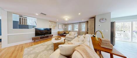 A spacious living room with a large flat-screen TV, a sofa set, a dining area, and a view of the outdoors.