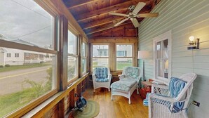 Una habitación con techo de madera, ventilador de techo, sillas de mimbre, una lámpara y ventanas grandes.