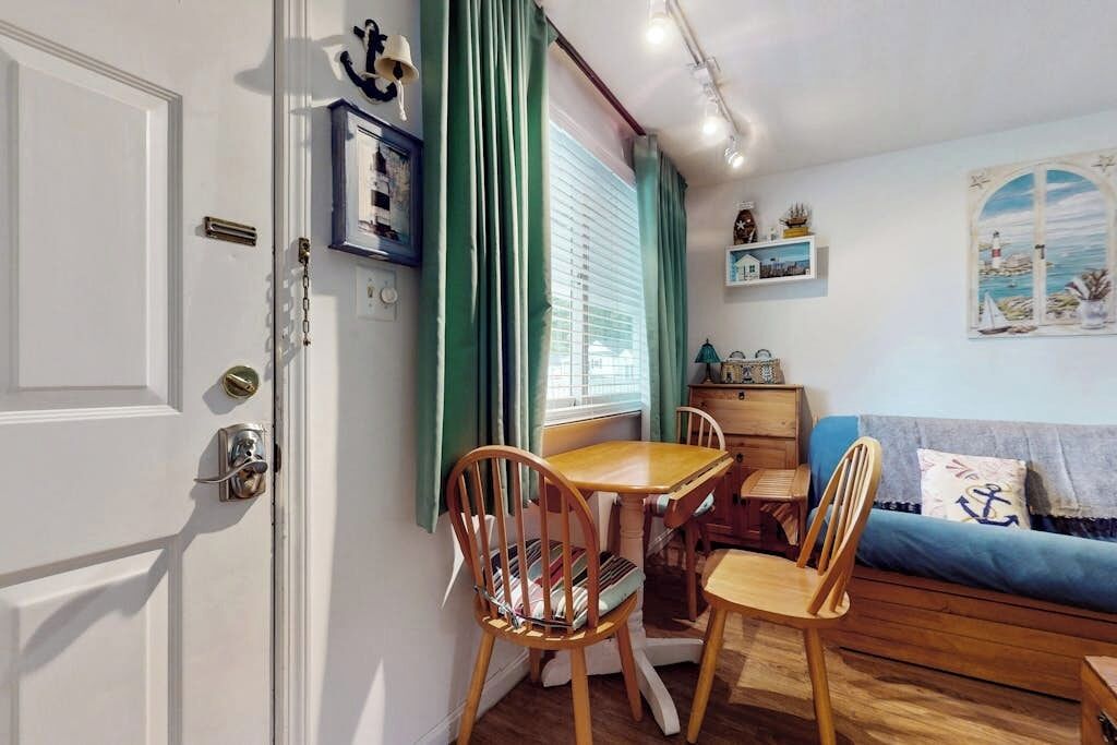 Une pièce comprenant une table à manger et des chaises, un canapé, une fenêtre avec des rideaux, une étagère fixée au mur ornée d’objets décoratifs et une porte avec une poignée.