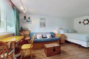 A compact room with a bed, a sofa, a dining table with chairs, a small cabinet, and a wooden chest.