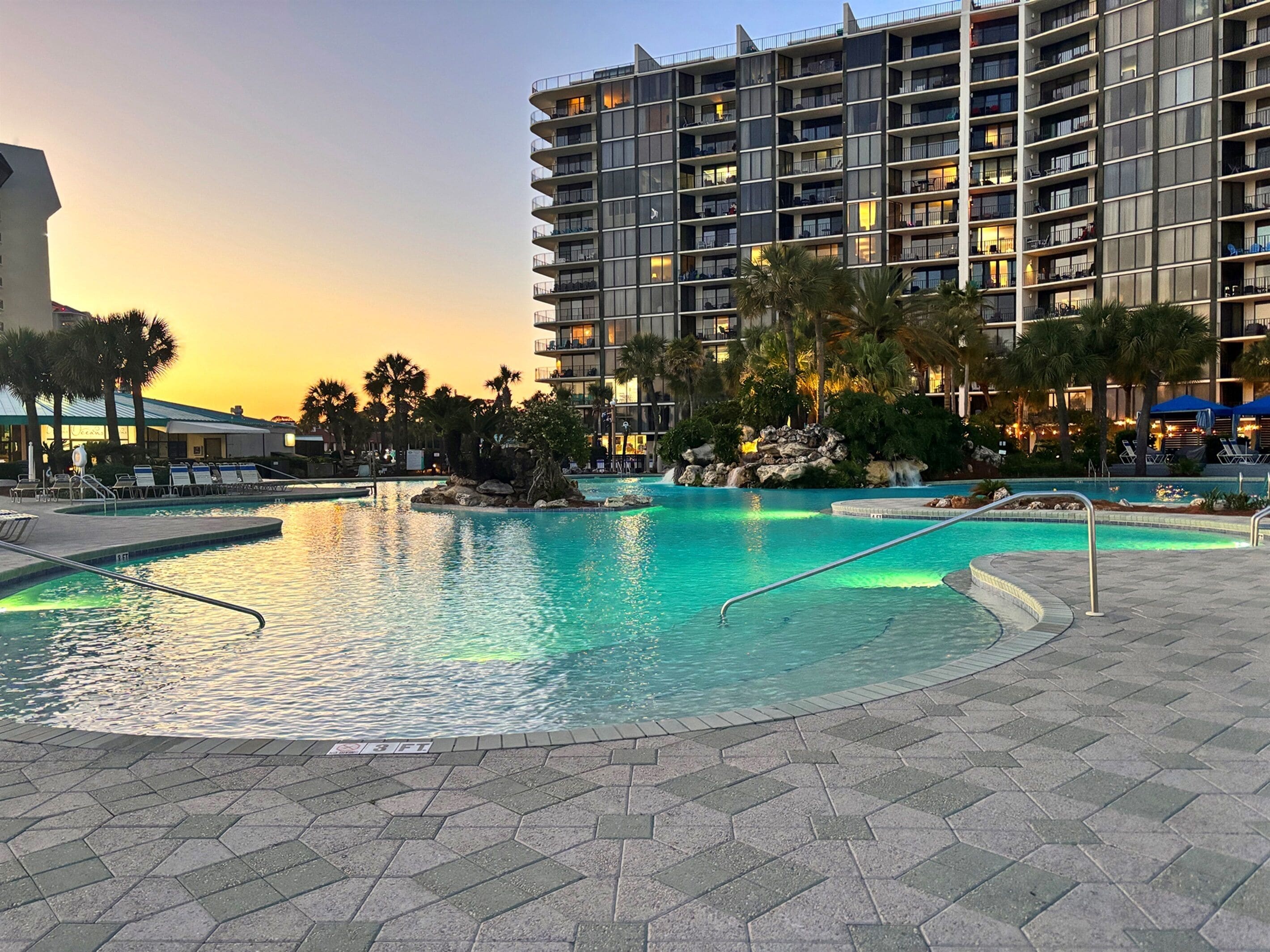 A hotel with a large outdoor swimming pool, palm trees, and a multi-story building with balconies.