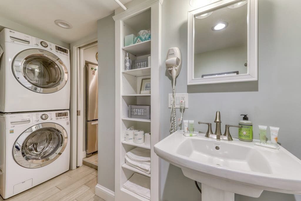 A laundry area with stacked washing machines, a drying machine, a mirror, and a sink with toiletries.