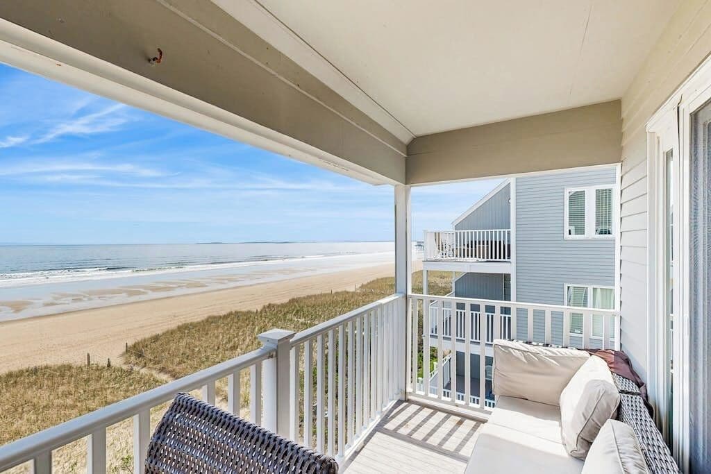 A balcony with a view of the beach, a white railing, and a sofa with cushions.