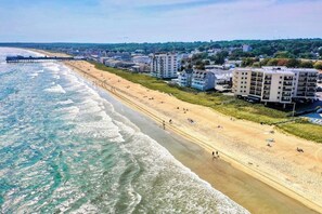 Eine Küstenlandschaft mit einem Strand und einer Pier, die sich ins Meer erstreckt, neben einer Reihe mehrstöckiger Gebäude.