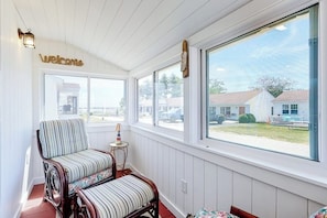 A sunroom with a 'Welcome' sign, a striped chair, a small table, and a view of houses outside.