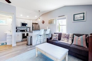 A modern kitchen with white cabinets, a stainless steel refrigerator, and a microwave. There is a dining area with a white table and chairs. A dark sofa with patterned cushions is in the living room area.
