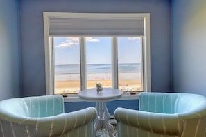 A room with a view of the beach, two upholstered chairs, a small table, and a window with blinds.