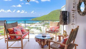 Uma varanda com móveis de vime, uma mesa de vidro redonda e vista para o oceano e as colinas.
