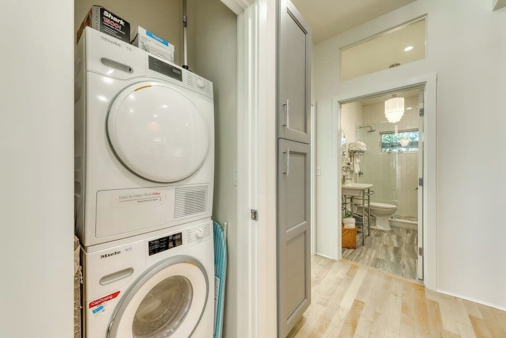 Un área de lavandería con lavadoras y secadoras apiladas, una puerta que da a un baño con ducha y lavamanos.