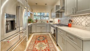 Une cuisine moderne dotée d’appareils électroménagers en acier inoxydable, de meubles de rangement gris, d’un tapis à motifs et d’une fenêtre laissant entrer la lumière naturelle.