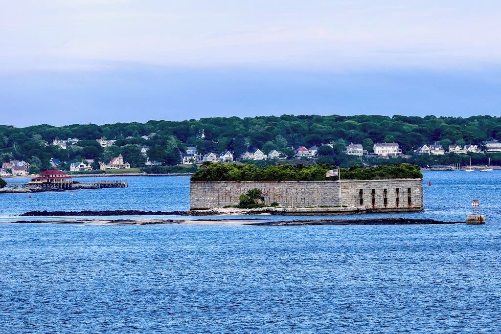 Un muelle de piedra que se adentra en una masa de agua, con un pequeño edificio en la parte superior, rodeado de una colina boscosa y edificios residenciales al fondo.