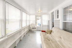 A room with a white ceiling, large windows with blinds, a white chair, a beige sofa, and a door leading outside. - Coastal Cottage w/ Enclosed Front Porch, Pond View – By Beach & Bike Path (Old Orchard Beach)