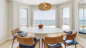 A round table with six wicker chairs, a hanging light fixture, and a view of the ocean through large windows. - Coastal gem on the water with beach access, gorgeous views & spacious layout (Wells)