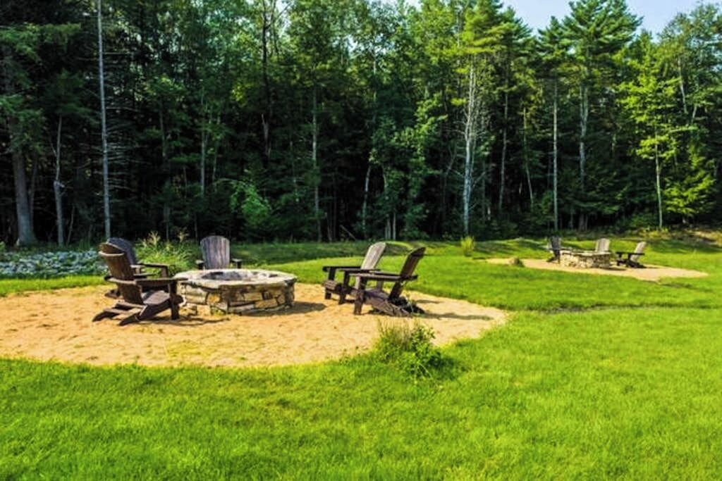 A campsite with a stone fire pit surrounded by Adirondack chairs, set in a grassy area with dense forest in the background.