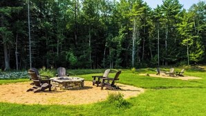 Un campeggio con un focolare in pietra circondato da poltrone Adirondack, immerso in un'area erbosa con una fitta foresta sullo sfondo.